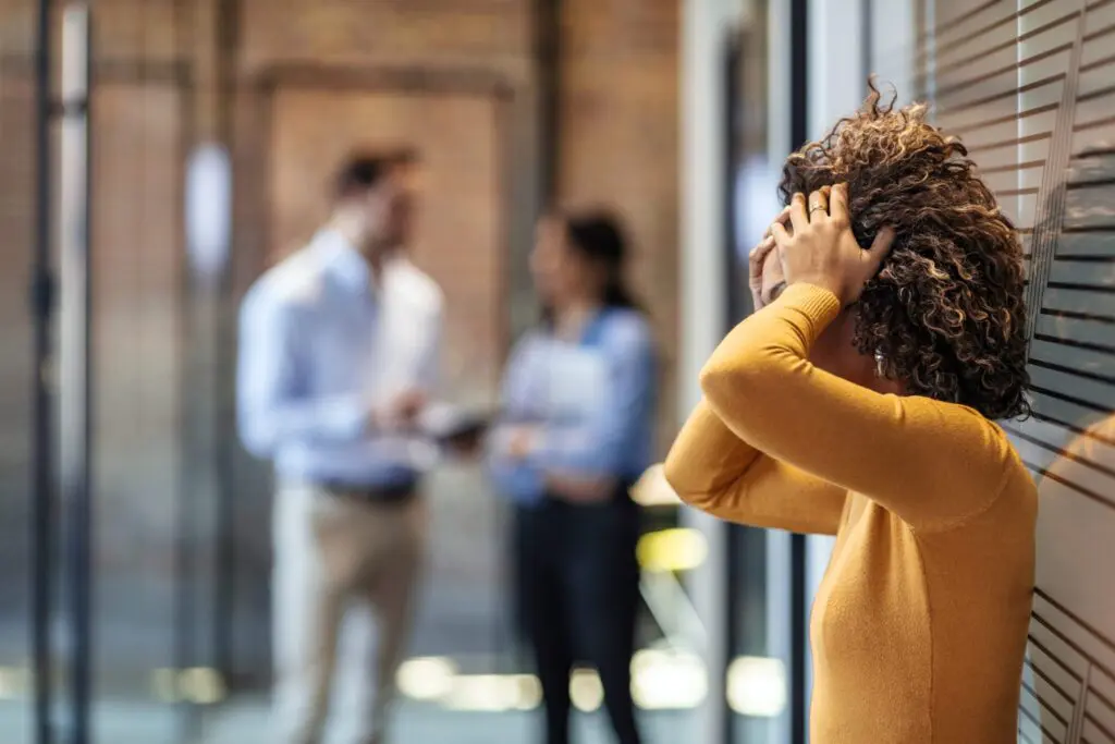 A stressed woman holding her head while two of her colleagues have a discussion in the background