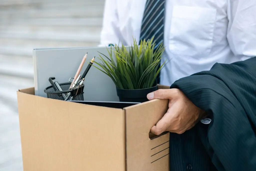 A businessman carrying a cardboard box filled with office supplies and a plant