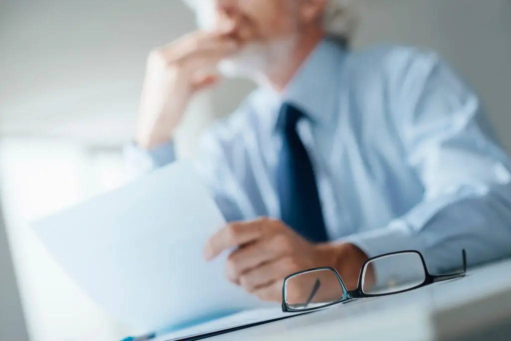 Man sitting at a desk with eyeglasses rubbing chin and thinking while holding a paper