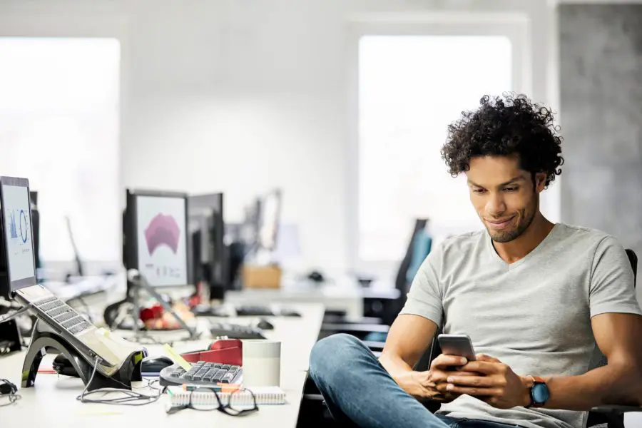 A man sits at his desk in a bright office, glancing at his phone amidst computers and office supplies. He's likely gathering tips for engaging millennials in the modern workplace.