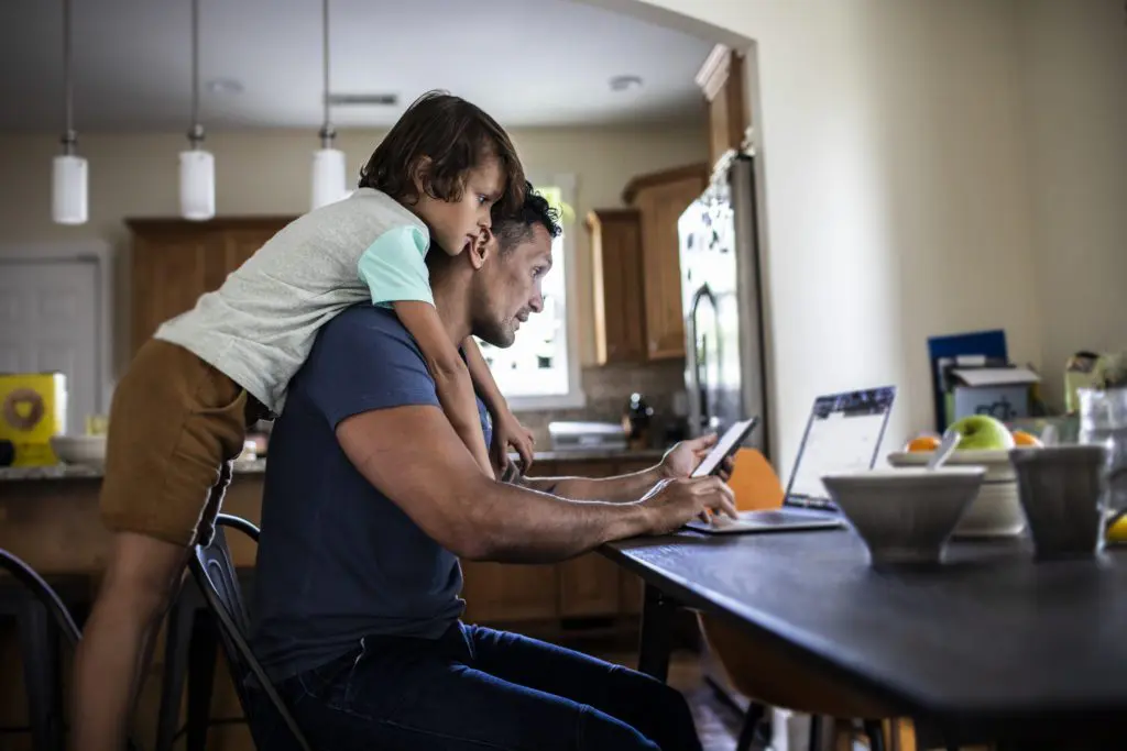 A young boy hanging over his dad's shoulders while he works on a computer at kitchen table