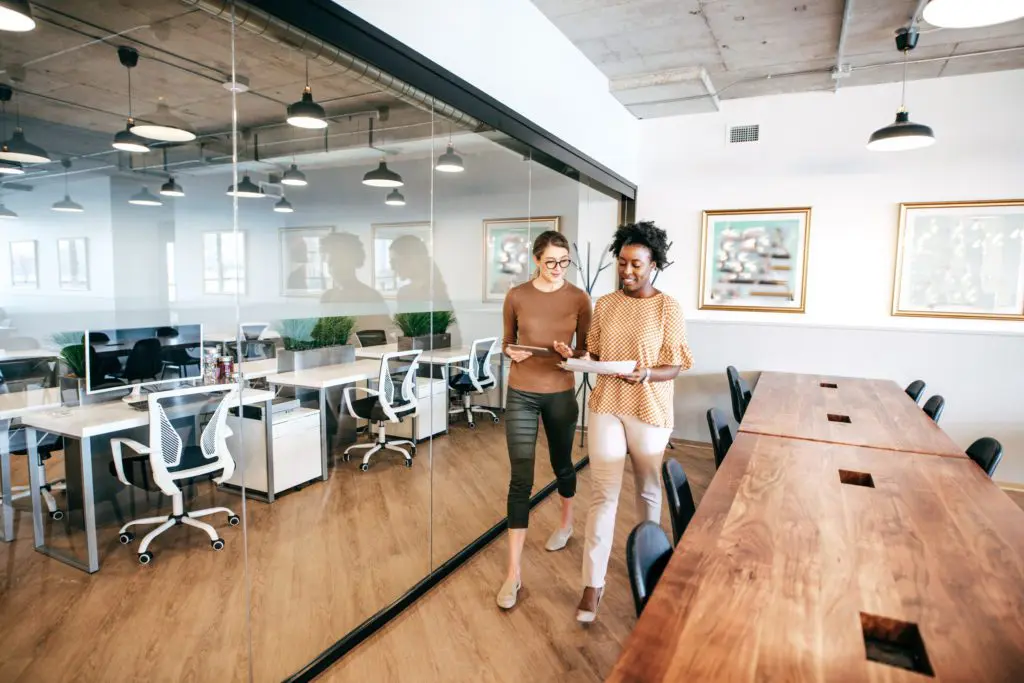 Two businesswomen walking and talking with tablet and papers in modern office