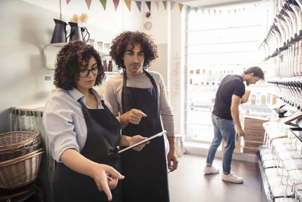 Two associates in aprons in shop pointing to products and looking at a clipboard