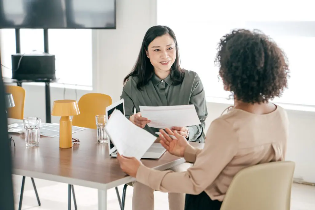 Two businesswomen sitting at a table talking and looking at paperwork