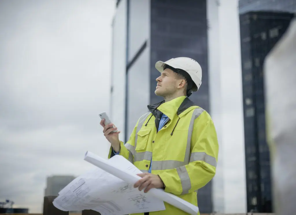 Worker in protective gear looking around a construction site with a phone and plans in his hands