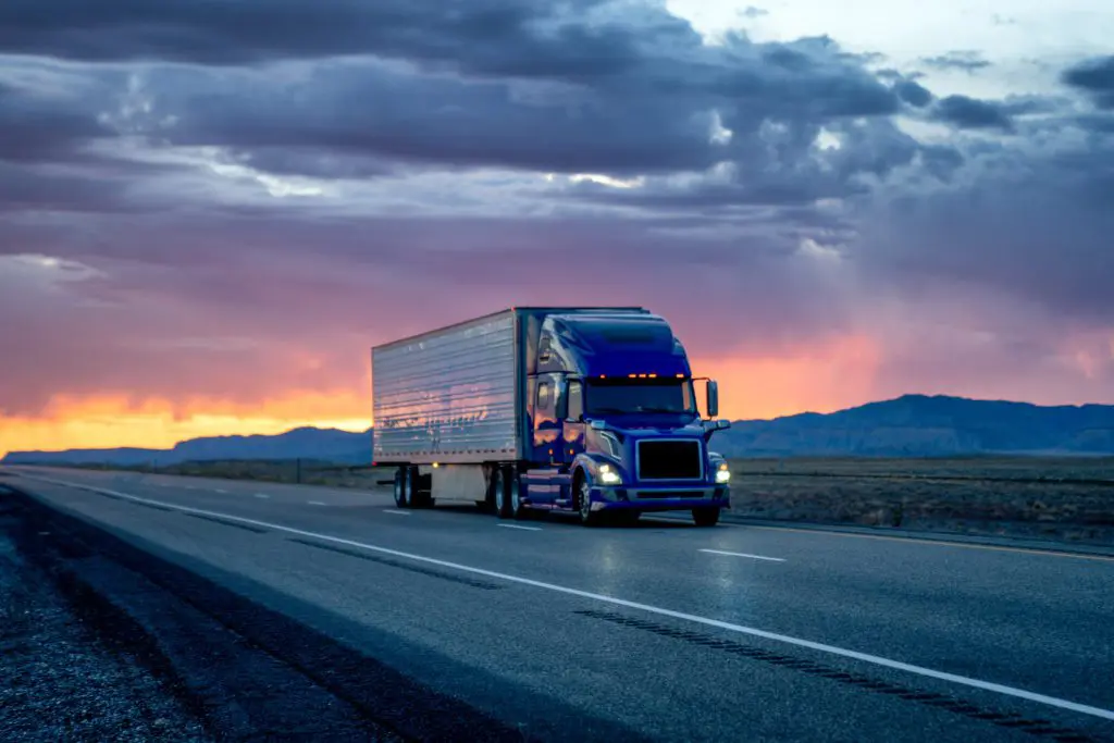 Semitruck driving down an open road surrounded by trees and a pretty sunset