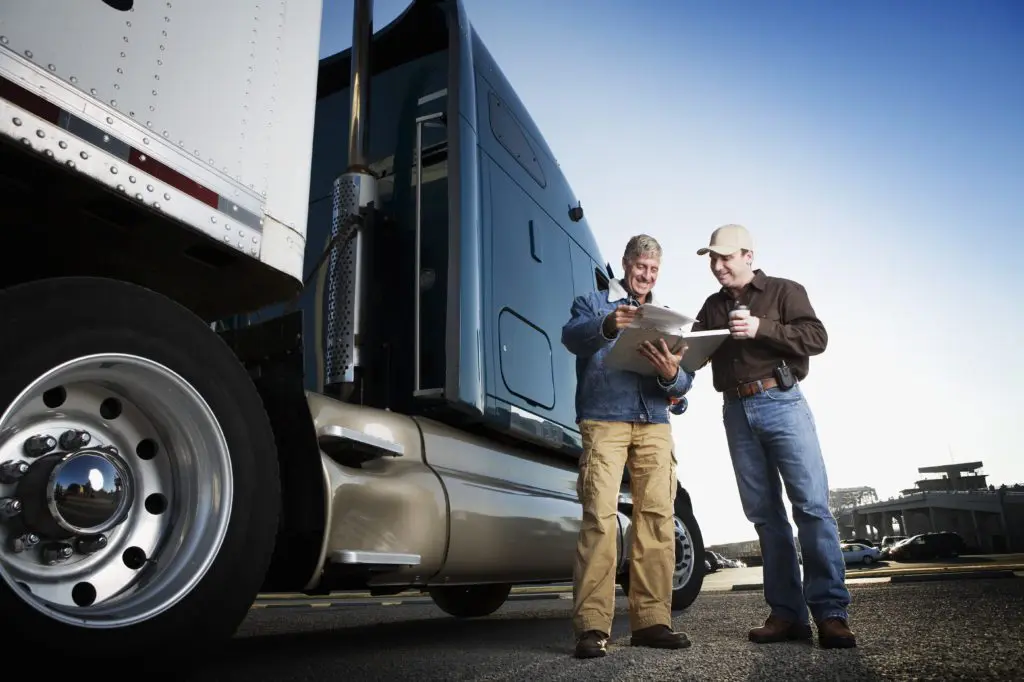 Two men next to a semi truck smiling and filling out paperwork on a clipboard