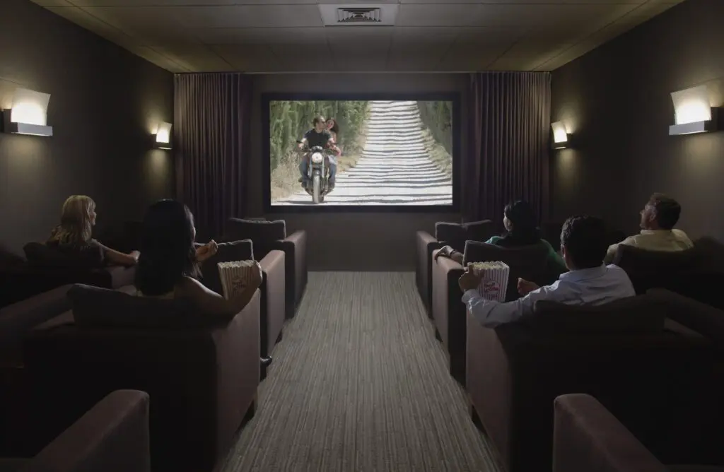 In a small, dimly lit home theater, people watch a movie featuring a motorcycle scene