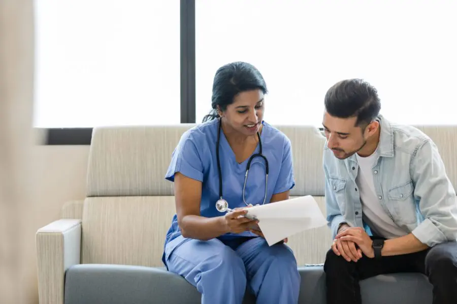 The young man and female doctor sit on the hospital room couch and review the medical chart together.