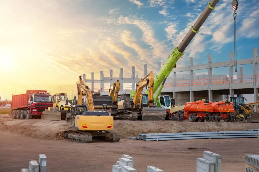 Many different multicolored colorful heavy industrial machinery equipment at construction site parking area against warehouse building city infrastructure development. Commercial vehicles rental sale.