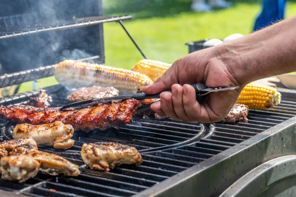 Hand using tongs to flip ribs on a grill surrounded by corn and chicken