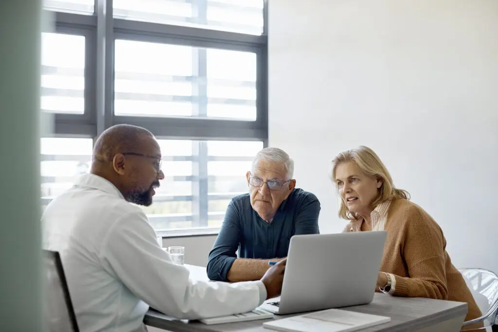 Male doctor explaining senior couple over laptop. Elderly man and woman are listening to medical expertise. They are sitting at desk in hospital.