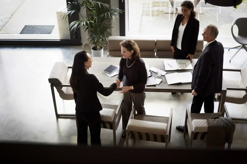 Group of professional associates around a table shaking hands and talking in a modern office