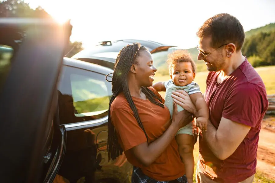 Smiling parents holding their baby near a car in a sunny outdoor setting