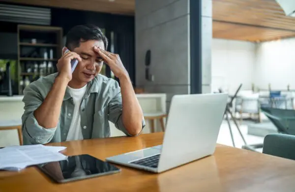 A businessman appears stressed while talking on the phone at his desk in a modern office setting. He holds his head, surrounded by documents, a laptop, and a digital tablet.