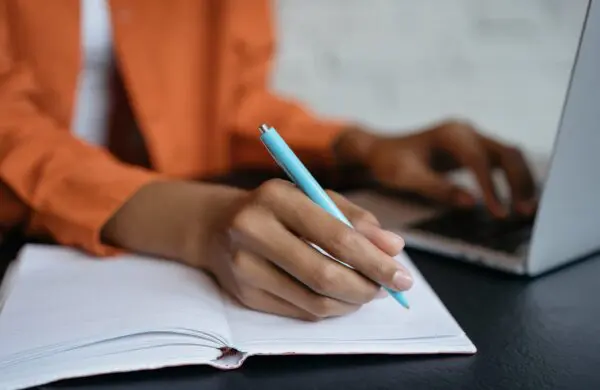 A HR professional writes in a notebook with a pen while typing on a laptop, writing a description for a job posting