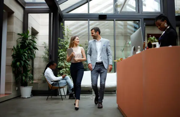 Two people walk together in a modern office space with large windows and plants, embracing sustainability in the workplace. Another person sits in the background, while a woman stands at a reception desk.
