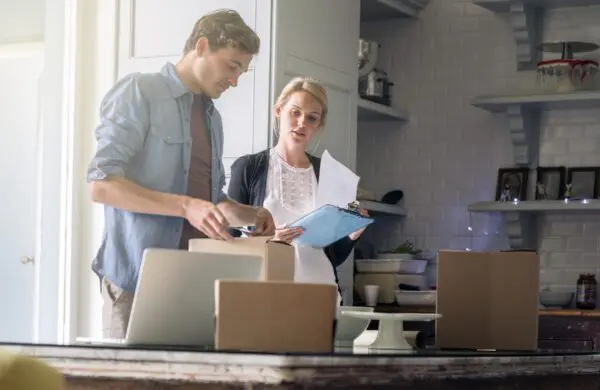 Amidst the backdrop of shelves and kitchenware, two people are busy in the kitchen. One, with a clipboard and papers, meticulously manages the home inventory while the other efficiently packs items into cardboard boxes.