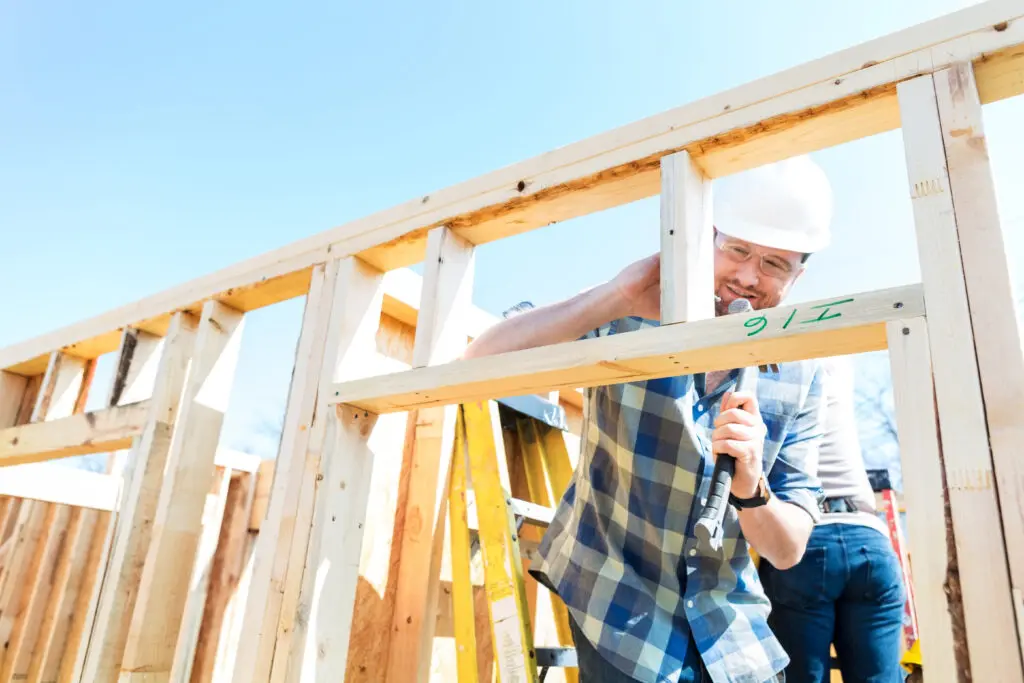Mid-adult construction worker or building contractor wearing safety glasses and a hard hat uses a hammer to build house frame