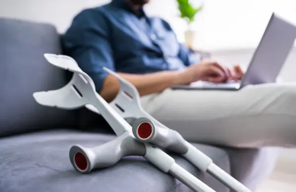 Man with a leg injury sits on a sofa working on his laptop with crutches laid beside him