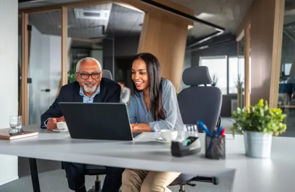 A senior man in professional attire works with a businesswoman at a laptop in a modern office