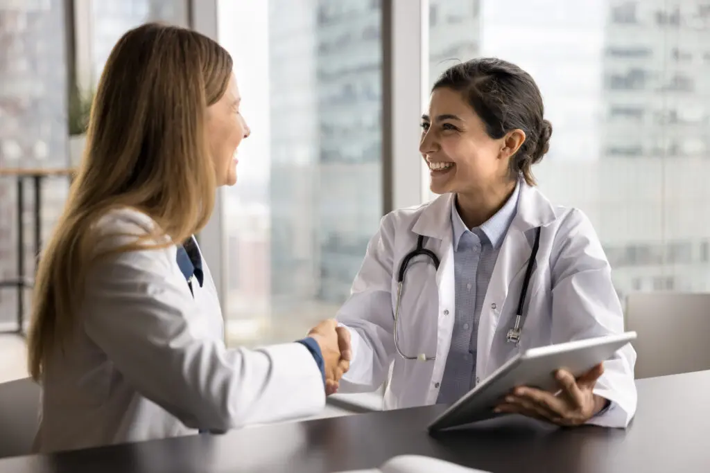 Two female doctors shake hands seated across table, holding tablet with medical information