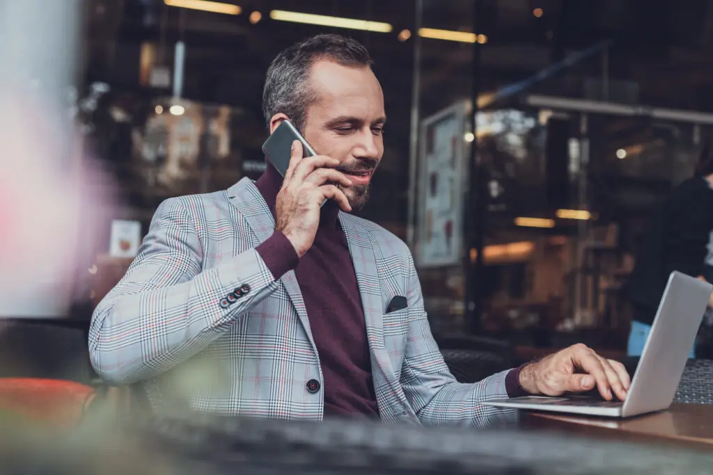 Young man sitting at a table and looking at the screen of his modern laptop while having a phone conversation