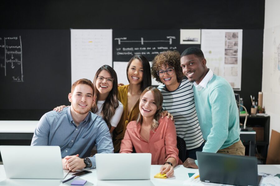 A group of smiling business professionals gathered around their laptops in the office