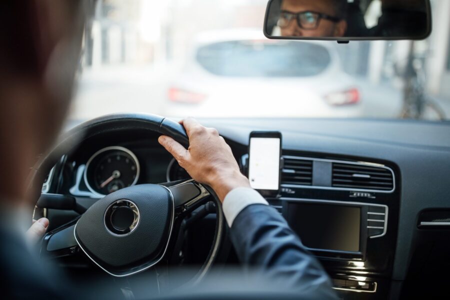A man in a suit driving a car while his reflection is visible in the rearview mirror