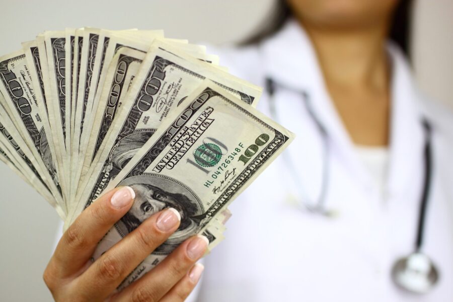 Health care worker in a white coat holding a fan of $100 bills with a stethoscope around their neck