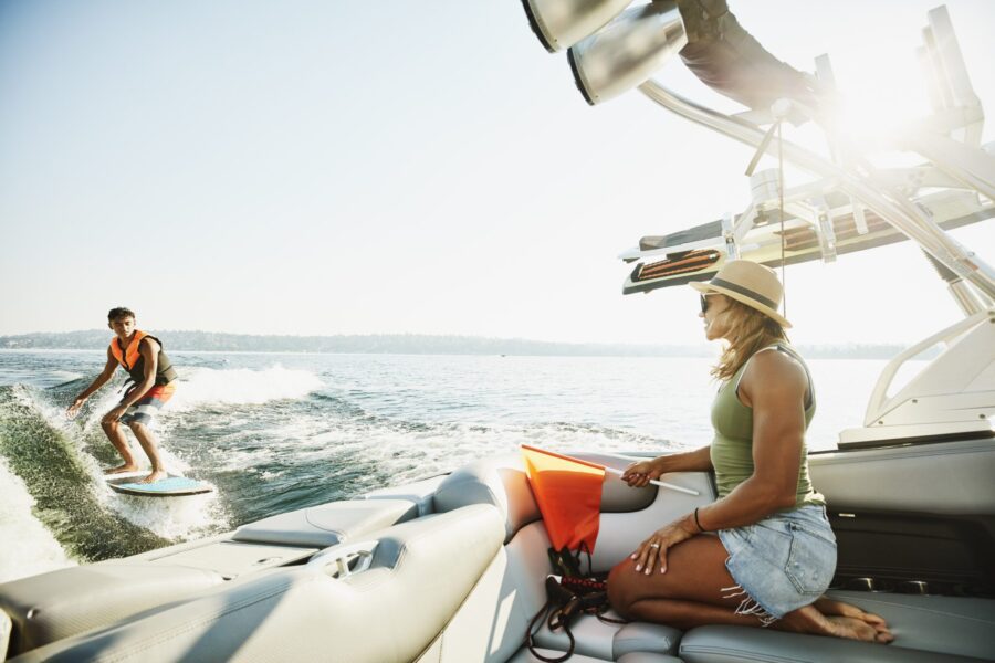 A woman sitting on a boat watching a man wake surf while they enjoy a sunny day on the water