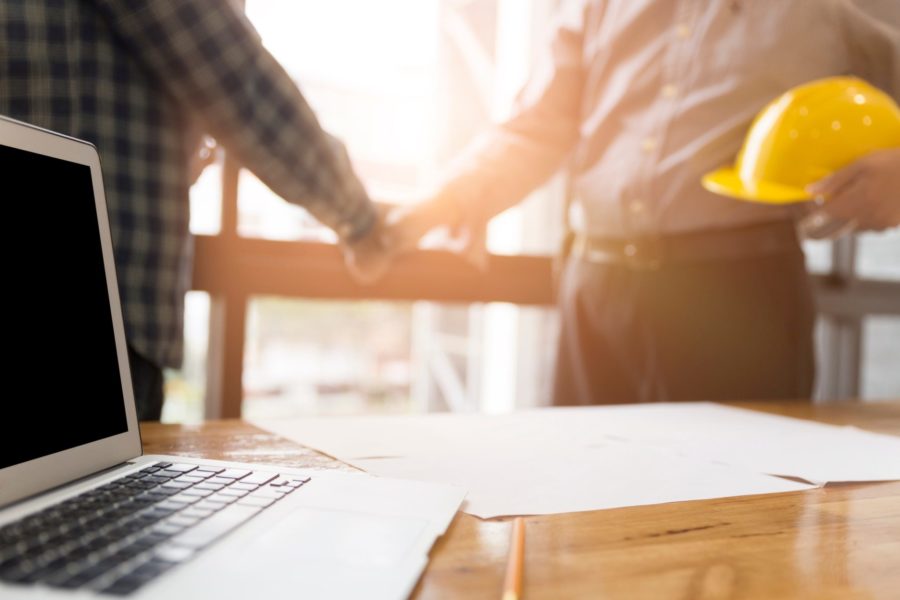 A laptop and architectural plans lie on a desk, while two people shake hands in the background, one with a yellow hard hat. Their discussion likely touches on Texas anti-indemnity laws and construction contract risk management.