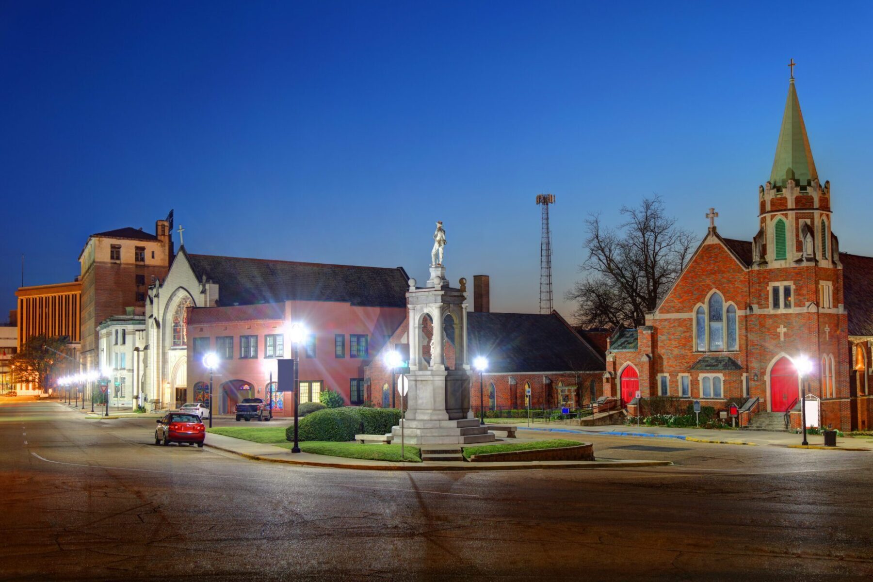 Historic buildings and a church with a tall steeple and red doors are illuminated at dusk in a town square in Texarkana