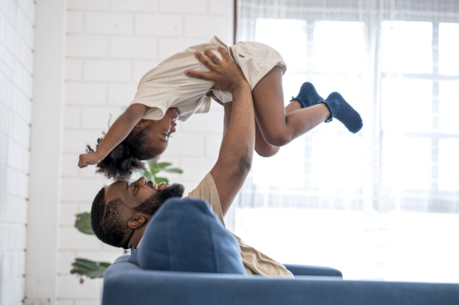 African Father Picking Up Daughter In The Air, Face To Face. They Are Having Fun In Weekend At Home.