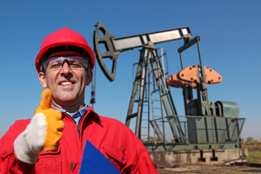 A smiling oilfield worker giving a thumbs up with an oil pump visible in the background