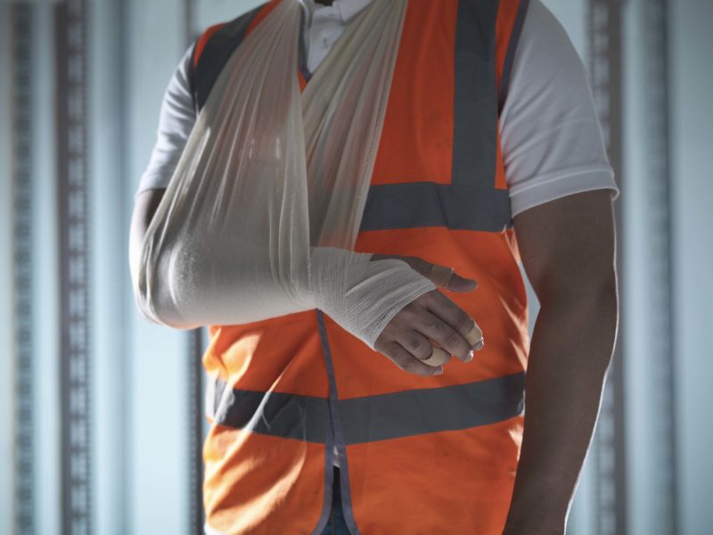 A person wearing an orange safety vest with one arm in a sling and bandaged hand is standing indoors, seemingly acclimating to the challenging experience of their injury.