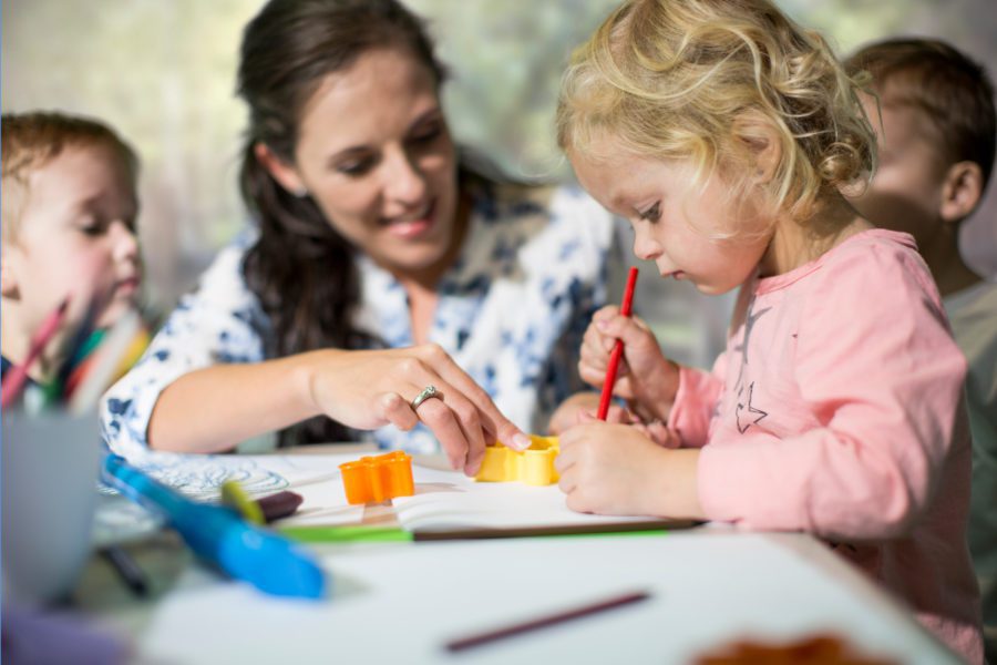 A woman sits with young children at a table. She and a child focus on crafting with colored paper and pencils.