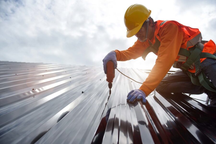 A construction worker in safety gear using a drill to install metal roofing