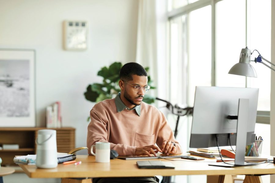 A man in a peach sweater works diligently at his computer, running his home-based business from a well-lit office adorned with plants, books, and a desk lamp.