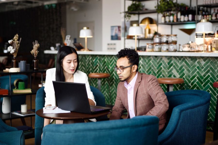 Two people sit in a stylish cafe, deeply engaged in a discussion about errors and omissions while looking at a laptop. A bar with a green tile backdrop adds to the vibrant atmosphere in the background.