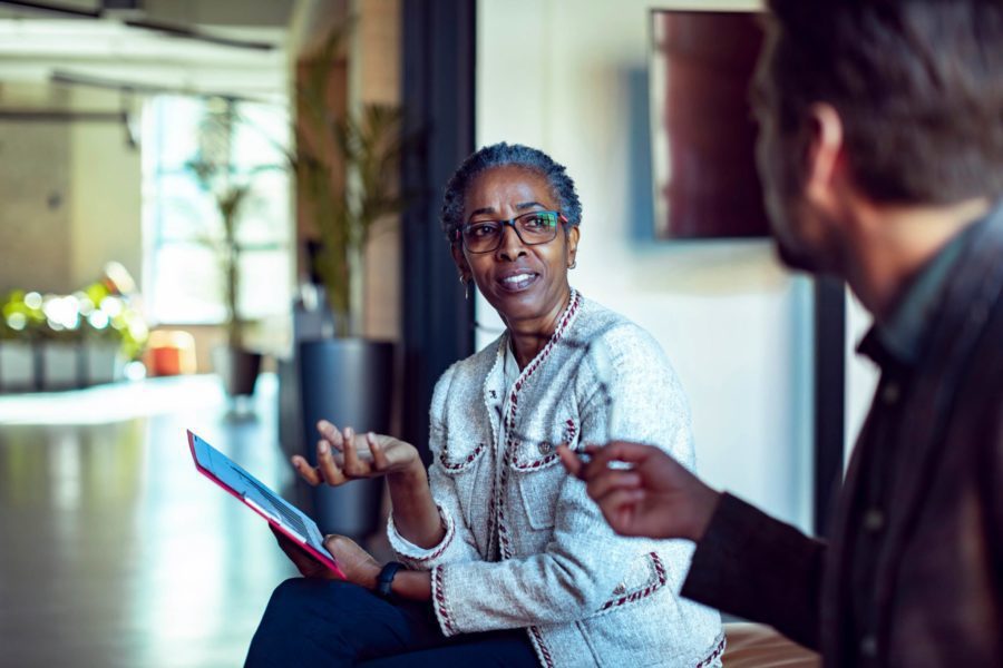 Two people are engaged in a conversation indoors. One, embodying the traits of a great boss, is holding a tablet and gesturing animatedly as they speak, while the other listens attentively.