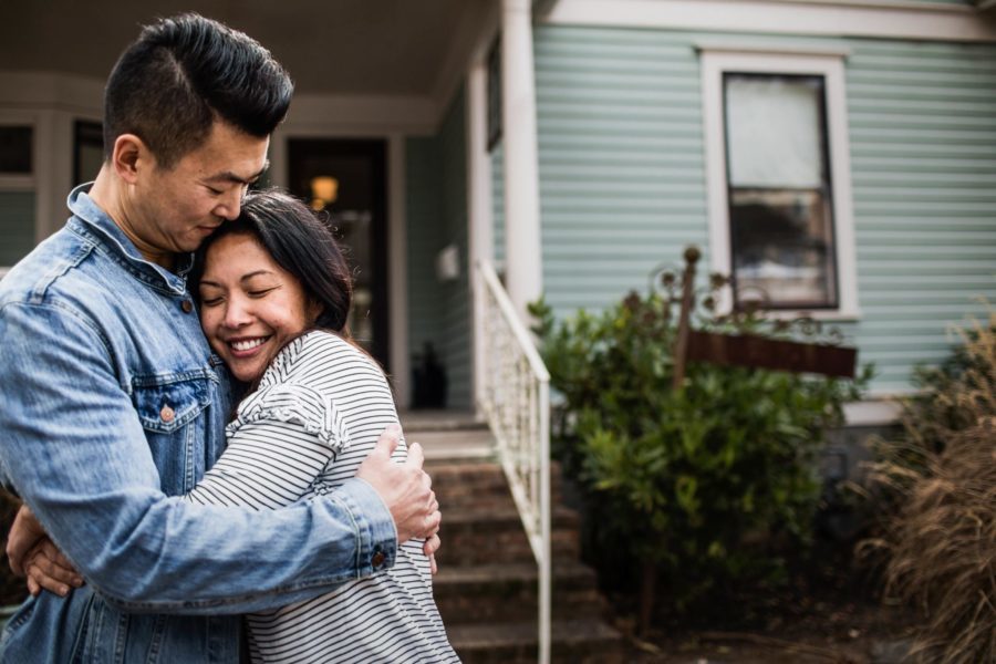 A couple embraces happily outside a house with a light blue exterior and white trim, feeling secure with mortgage protection. A staircase leads up to the entrance beside them.