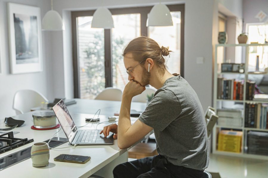 A person with tied-back hair sits at a kitchen table, exploring side hustles on a laptop while wearing earphones. Nearby, a smartphone and mug wait in the modern decor, where large windows and pendant lights add a contemporary touch.