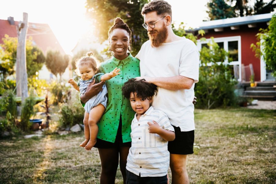 In a sunlit yard that feels like home, a family of four smiles brightly. The parents stand together with two young children, one lovingly held by the mother.