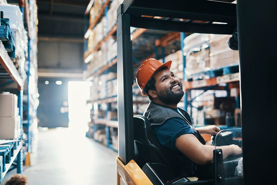 A person wearing an orange hard hat operates a forklift in a warehouse, surrounded by tall shelves filled with boxes, mindful of general liability as they navigate the busy workspace.
