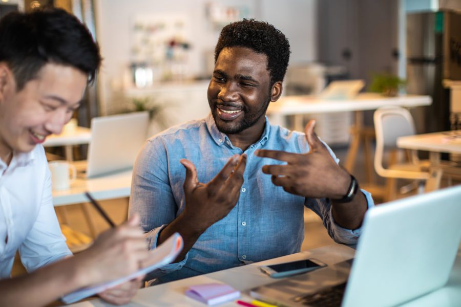 Two men are sitting at a desk in an office, smiling and talking about work in insurance. One takes notes while the other gestures. A laptop and smartphone rest on the desk, underpinning their discussion.