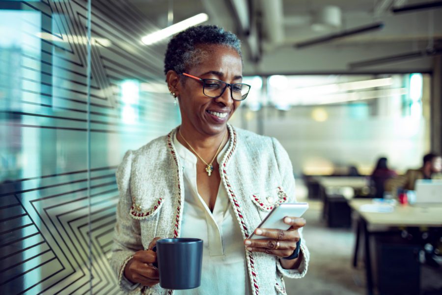 In a bright office, someone in a light-colored jacket smiles at their smartphone, clutching a mug. Perhaps they just mastered the transfer of business contacts from Android to iPhone, enhancing their connectivity and productivity.