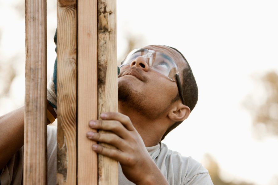 A person wearing safety glasses, who recently obtained their contractor's license, inspects and holds wooden beams, gazing upwards.