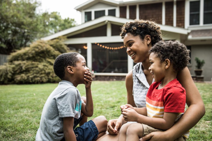 A woman sits on the grass with two boys, smiling and laughing, in a backyard setting. Behind them, a charming house stands quietly, its true worth known only to those exploring off-market gems.