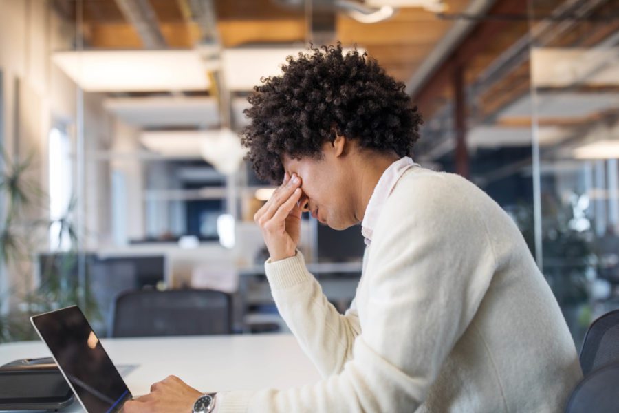 A person with curly hair sits at a desk in an office with glass walls and overhead lights, resting their head in one hand while looking at a laptop, perhaps pondering ways to promote stress relief amidst the daily grind.